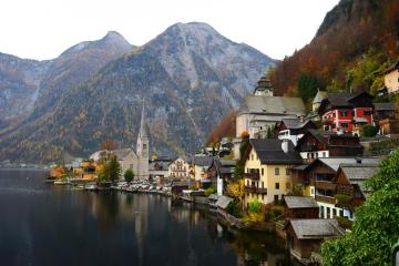 A picturesque village with traditional alpine houses and a prominent church steeple situated on the shore of a calm lake, backed by steep, forested mountains.