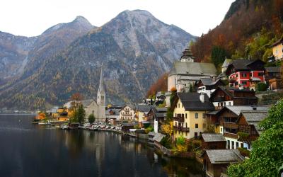 A picturesque village with traditional alpine houses and a prominent church steeple situated on the shore of a calm lake, backed by steep, forested mountains.