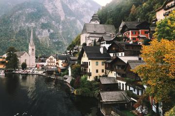The image shows the lakeside village of Hallstatt in Austria, featuring its iconic church spire, traditional alpine houses, and the Dachstein mountains in the background, with autumn foliage visible on the right.