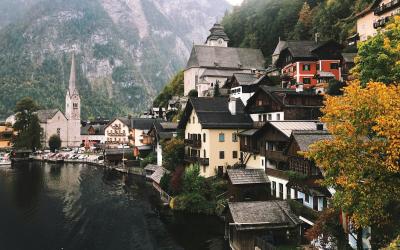 The image shows the lakeside village of Hallstatt in Austria, featuring its iconic church spire, traditional alpine houses, and the Dachstein mountains in the background, with autumn foliage visible on the right.