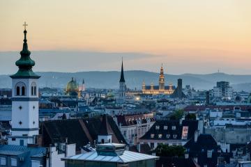 A panoramic cityscape at dusk showing a prominent white church tower with a green spire in the foreground, a golden-domed palace (Schönbrunn Palace) illuminated in the distance, and rolling hills on the horizon.
