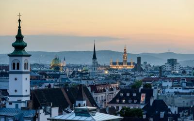 A panoramic cityscape at dusk showing a prominent white church tower with a green spire in the foreground, a golden-domed palace (Schönbrunn Palace) illuminated in the distance, and rolling hills on the horizon.