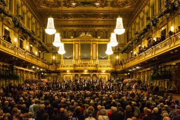 The image shows the interior of a grand concert hall with ornate gold decoration, multiple tiers of balconies, a large pipe organ on the stage wall, and a full orchestra performing for a seated audience.