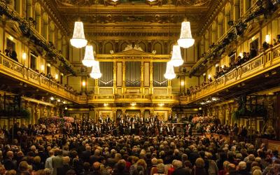 The image shows the interior of a grand concert hall with ornate gold decoration, multiple tiers of balconies, a large pipe organ on the stage wall, and a full orchestra performing for a seated audience.