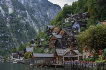 A cluster of traditional wooden houses with steep roofs and colorful shutters built on a steep hillside next to a calm lake, with a large forested mountain in the background; no visible signage or text.