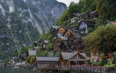 A cluster of traditional wooden houses with steep roofs and colorful shutters built on a steep hillside next to a calm lake, with a large forested mountain in the background; no visible signage or text.