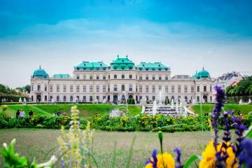 The image shows the Upper Belvedere Palace in Vienna, Austria, a grand Baroque palace with a distinctive green copper roof, set against a blue sky with manicured gardens and fountains in the foreground.