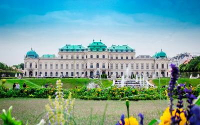 The image shows the Upper Belvedere Palace in Vienna, Austria, a grand Baroque palace with a distinctive green copper roof, set against a blue sky with manicured gardens and fountains in the foreground.