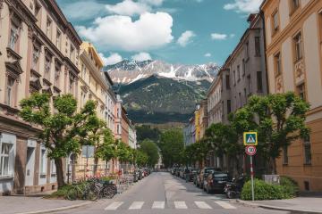 A street scene in a European city with multi-story buildings lining both sides, parked cars and bicycles along the curb, a pedestrian crosswalk, and a large snow-capped mountain visible at the end of the street under a partly cloudy sky.