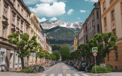 A street scene in a European city with multi-story buildings lining both sides, parked cars and bicycles along the curb, a pedestrian crosswalk, and a large snow-capped mountain visible at the end of the street under a partly cloudy sky.