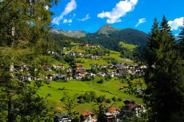 A scenic view of a small alpine village nestled in a green valley with traditional houses, surrounded by forested hills and a prominent mountain peak under a blue sky with scattered clouds.