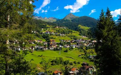 A scenic view of a small alpine village nestled in a green valley with traditional houses, surrounded by forested hills and a prominent mountain peak under a blue sky with scattered clouds.