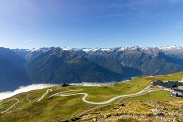 A high-angle landscape view of a winding mountain road (the Bramberg Pass or similar alpine route) with a grassy foreground, distant snow-capped peaks, and a small building complex on the right.