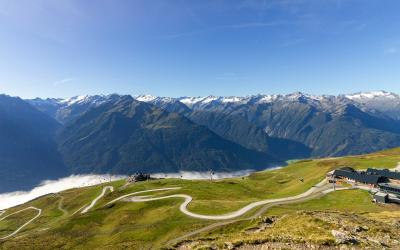 A high-angle landscape view of a winding mountain road (the Bramberg Pass or similar alpine route) with a grassy foreground, distant snow-capped peaks, and a small building complex on the right.