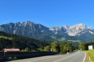 A paved two-lane road curves through a green valley with a prominent, rugged mountain range in the background under a clear blue sky; roadside signage is visible but illegible.