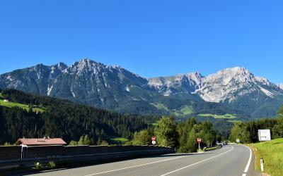 A paved two-lane road curves through a green valley with a prominent, rugged mountain range in the background under a clear blue sky; roadside signage is visible but illegible.