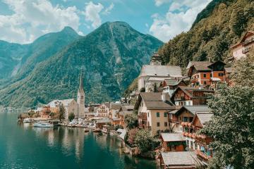 A scenic view of the village of Hallstatt, Austria, featuring its iconic church steeple, colorful houses along the shore of Lake Hallstatt, and the Dachstein mountains in the background, with a blurred foreground element.
