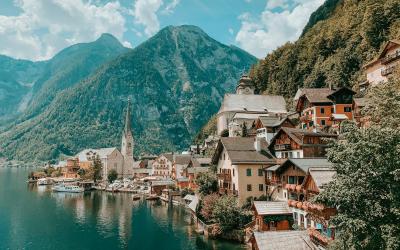 A scenic view of the village of Hallstatt, Austria, featuring its iconic church steeple, colorful houses along the shore of Lake Hallstatt, and the Dachstein mountains in the background, with a blurred foreground element.