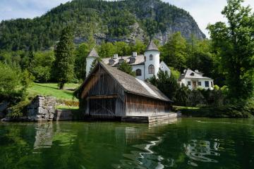 A wooden boathouse on a lake with a white church and houses on the shore, backed by a steep, forested limestone mountain.
