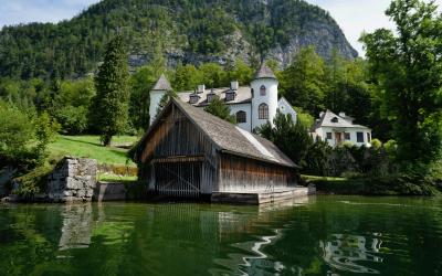 A wooden boathouse on a lake with a white church and houses on the shore, backed by a steep, forested limestone mountain.