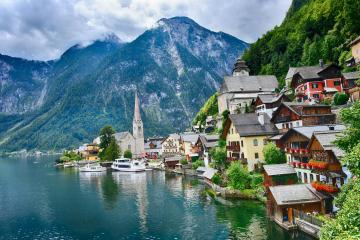 A scenic view of the village of Hallstatt on the shore of Lake Hallstatt in Austria, featuring traditional houses, a church steeple, mountains, and boats on the water.