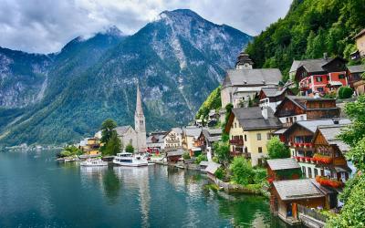 A scenic view of the village of Hallstatt on the shore of Lake Hallstatt in Austria, featuring traditional houses, a church steeple, mountains, and boats on the water.
