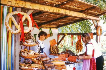 A rustic wooden sales booth at an outdoor market, featuring a large pretzel sign, hanging sausages, and a display of various breads and baked goods, with two women working behind the counter.