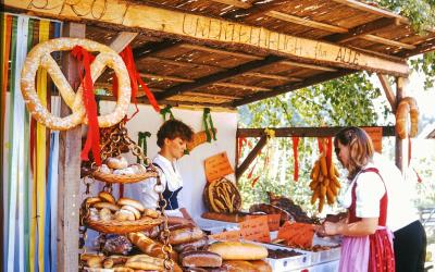 A rustic wooden sales booth at an outdoor market, featuring a large pretzel sign, hanging sausages, and a display of various breads and baked goods, with two women working behind the counter.