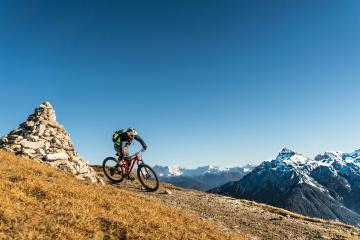 A mountain biker rides along a high-altitude ridge trail with a distinctive stone cairn on the left and snow-capped Alpine peaks in the background under a clear blue sky.