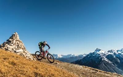 A mountain biker rides along a high-altitude ridge trail with a distinctive stone cairn on the left and snow-capped Alpine peaks in the background under a clear blue sky.