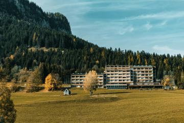 A large, multi-story institutional building complex situated in a grassy valley with a steep, forested mountain slope rising behind it and a few scattered trees in the foreground.