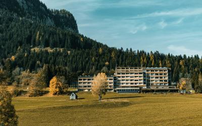 A large, multi-story institutional building complex situated in a grassy valley with a steep, forested mountain slope rising behind it and a few scattered trees in the foreground.