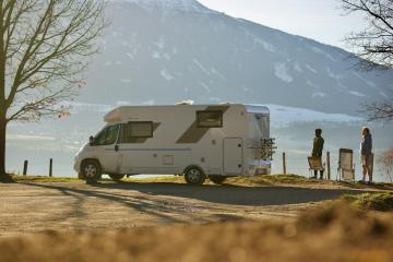 A white motorhome is parked on a dirt or gravel roadside area next to a body of water, with two people standing nearby and a large mountain in the background.