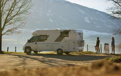 A white motorhome is parked on a dirt or gravel roadside area next to a body of water, with two people standing nearby and a large mountain in the background.