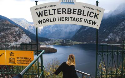 A woman stands on a viewing platform overlooking a lake and mountains, with a large sign reading "WELTERBEBLICK WORLD HERITAGE VIEW" and "ABSEITSPLATFORM / VIEWING PLATFORM".