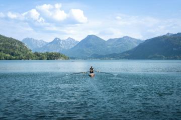 A person in a rowing shell is on a large lake with forested hills and mountains in the background under a partly cloudy sky.