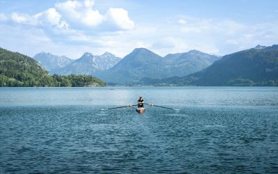 A person in a rowing shell is on a large lake with forested hills and mountains in the background under a partly cloudy sky.