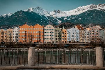 A row of colorful multi-story residential buildings along a riverbank with a metal railing in the foreground and snow-capped mountains in the background.