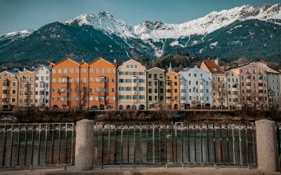 A row of colorful multi-story residential buildings along a riverbank with a metal railing in the foreground and snow-capped mountains in the background.