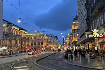 A wide European city street at dusk with tram tracks, historic architecture, overhead wires, and a "BISTRO" sign on the right.