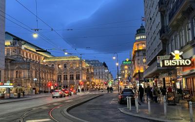 A wide European city street at dusk with tram tracks, historic architecture, overhead wires, and a "BISTRO" sign on the right.