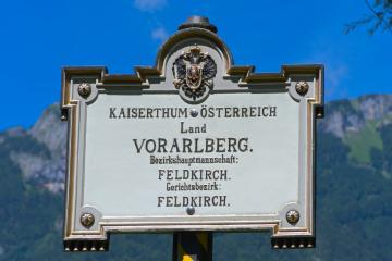 A vintage-style metal boundary sign reading "KAISERTHUM ÖSTERREICH Land VORARLBERG. Bezirksmannschaft: FELDKIRCH. Gerichtsbezirk: FELDKIRCH." with the Habsburg double-headed eagle emblem, set against a mountainous backdrop under a blue sky.