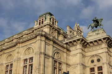 The image shows the upper facade and roofline of a grand, ornate stone building with classical architectural details, including arched windows, decorative cornices, and a prominent equestrian statue on the right; the sky is blue with scattered clo