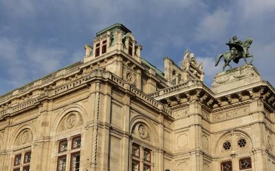 The image shows the upper facade and roofline of a grand, ornate stone building with classical architectural details, including arched windows, decorative cornices, and a prominent equestrian statue on the right; the sky is blue with scattered clo