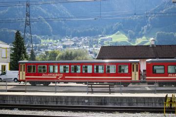 A red passenger train car with gold stripes and "ZILLERTALBAHN" written on the side, parked at a station platform with mountains and a village in the background.