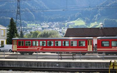 A red passenger train car with gold stripes and "ZILLERTALBAHN" written on the side, parked at a station platform with mountains and a village in the background.