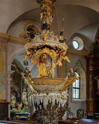 An ornate Baroque pulpit inside a church, featuring a boat-shaped sounding board with a net, a central painting of Jesus, and a column topped by a statue of Christ.