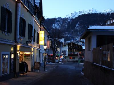 A street scene in a European town at dusk with snow-capped mountains in the background, featuring illuminated shop signs including one for "Maria" and another vertical sign reading "SCHLADMING".