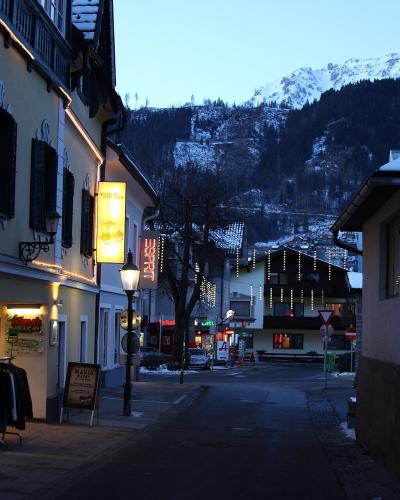 A street scene in a European town at dusk with snow-capped mountains in the background, featuring illuminated shop signs including one for "Maria" and another vertical sign reading "SCHLADMING".