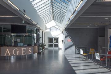 A modern office lobby with a reception desk displaying the letters "AGES", a glass skylight ceiling, and a waiting area with colorful chairs.